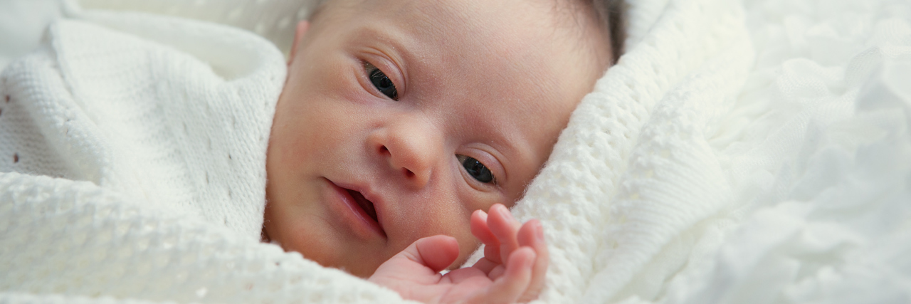 What Every Parent Facing a New Down Syndrome Diagnosis Should Know A baby with Down syndrome cuddled up in a white blanket