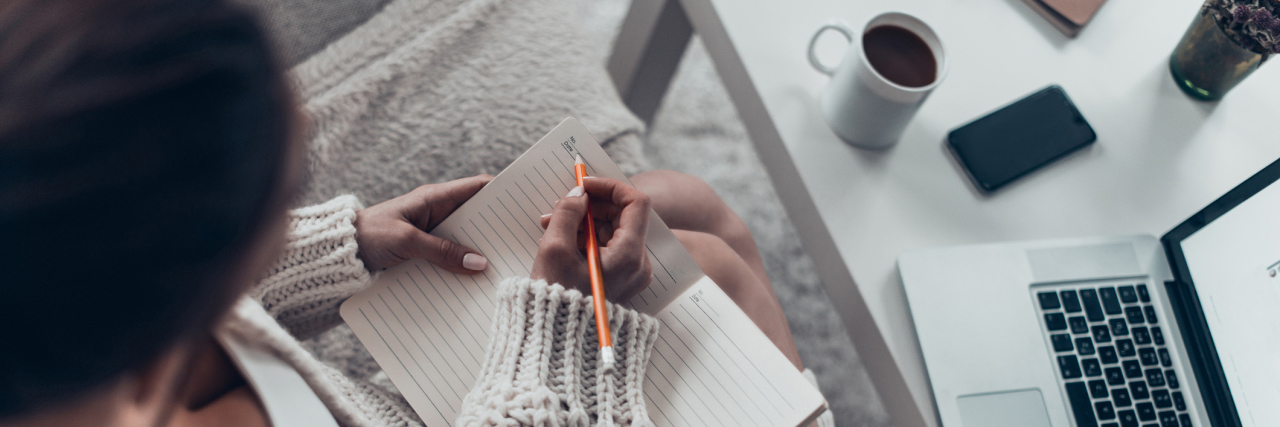 8 Ways You Can Use a Bullet Journal for Chronic Illness A woman sitting ny her computer and a cup of coffee, while journaling.