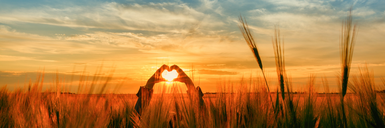 How I'm Expressing Love as My Illness Progresses A woman standing in a flied, making a heart shape with her hands as the sun is setting.