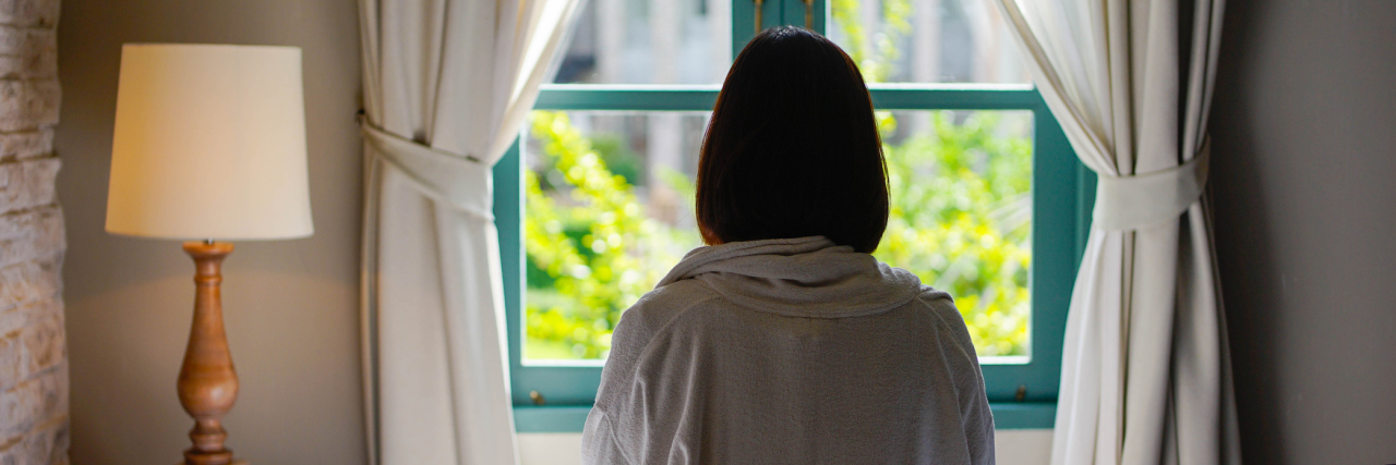 A picture of a woman sitting on her bed, looking out her window.