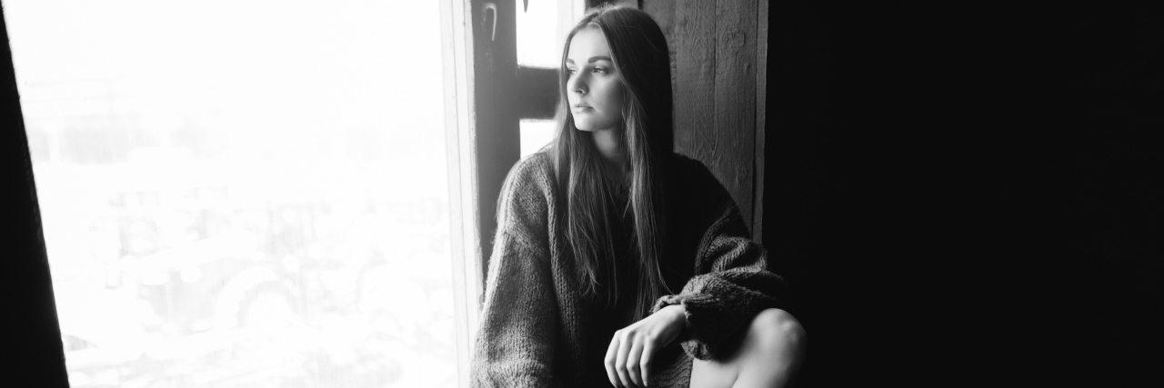 Why It's Hard to Get Anything Done When You're in Pain A black and white image of a woman sitting on a window cill, looking out the window.