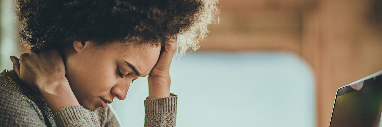 16 'Triggers' That Can Exacerbate Migraine Symptoms Black woman holding her head and neck in front of laptop
