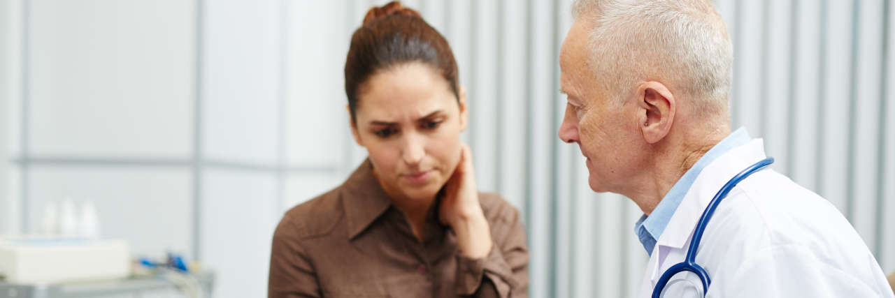 Things Doctors Often Say to Women, but Not Men Sad nervous young woman touching neck while reading her medical card shown by senior doctor in lab coat in modern office