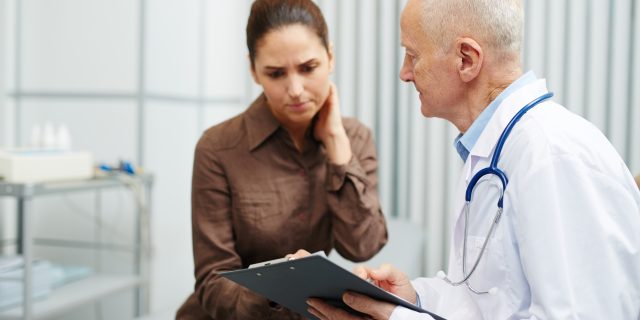 Sad nervous young woman touching neck while reading her medical card shown by senior doctor in lab coat in modern office