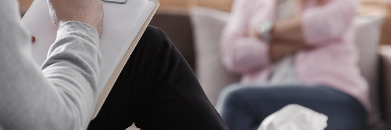 Back view of psychotherapist writing notes, assessing patient's health and giving diagnosis to a woman sitting on a couch in the blurred background during counseling session