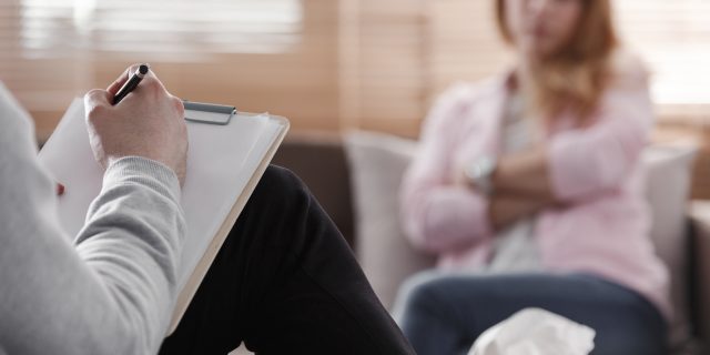 Back view of psychotherapist writing notes, assessing patient's health and giving diagnosis to a woman sitting on a couch in the blurred background during counseling session