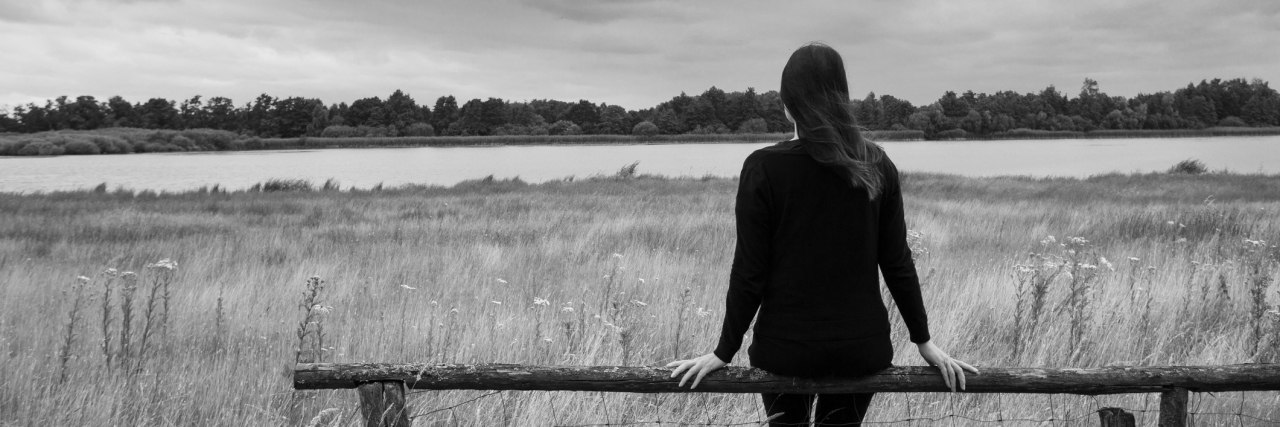 The Questions I Ask When People Refuse to Believe ME Exists A woman sitting alone on a bench, facing a field with a dark cloud above it.