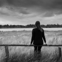 A woman sitting alone on a bench, facing a field with a dark cloud above it.