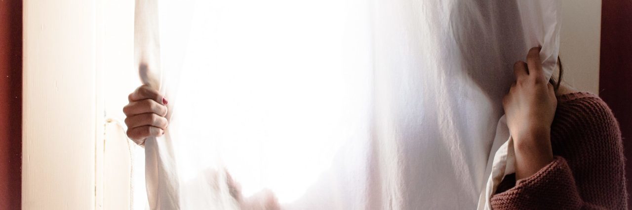 The Daily Cycle of Someone With Depression woman mostly hiding behind white curtain with only hands and shoulder visible