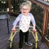 Adorable little girl using yellow walker, she is wearing glasses and giving a big smile at the camera.