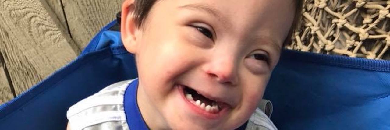 My Child With Down Syndrome Was Also Diagnosed With Cerebral Palsy Little boy with Down syndrome smiling at camera, he holds a water bottle with a red straw