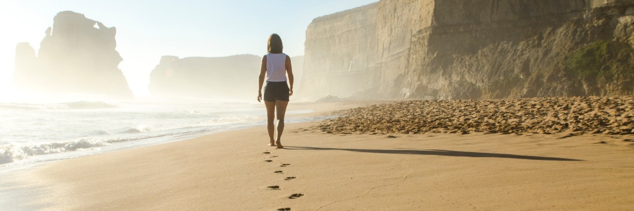 A woman walking alone on a bright summer beach, with her footsteps in the sand behind her as she walks away. 6 Unexpected Symptoms of Summer With Ehlers-Danlos Syndrome and POTS