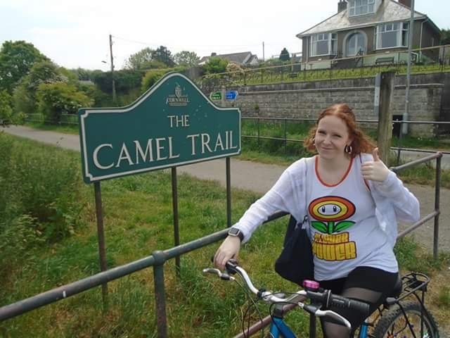 woman posing on her bike in front of a sign that says 'the camel trail'