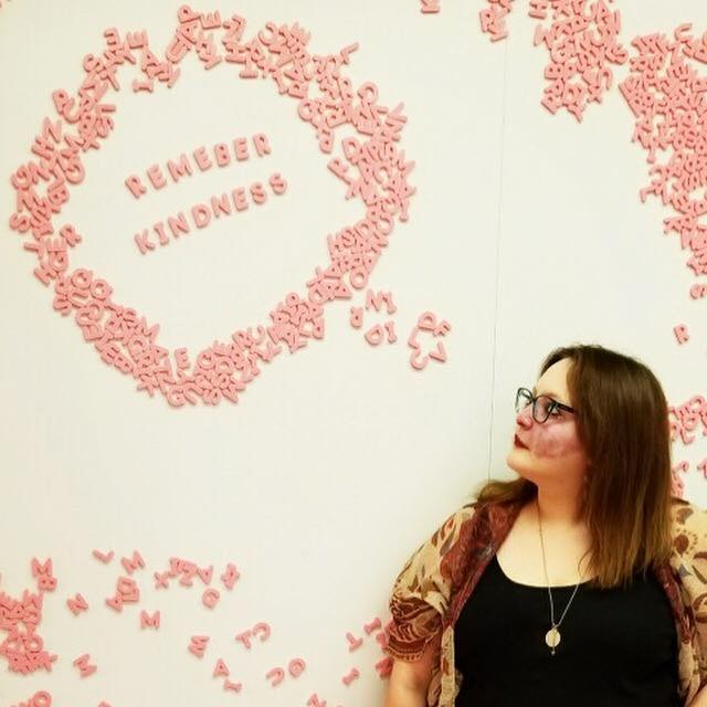 A picture of the writer standing in front of a magnetic wall covered with pink magnetic letters. Above Crystal, she created a speech bubble out of letters, and as she looks at it, it reads, "Remember kindness."