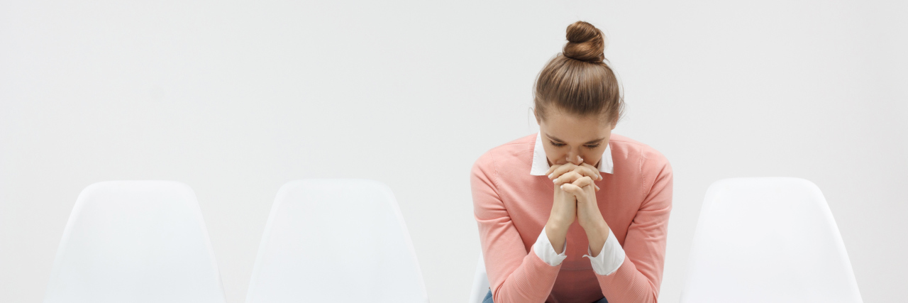 Why Being Undiagnosed Is Emotionally Draining Young woman sitting in the line or queue, looking stressed, putting hands together as if she is praying with closed eyes to overcome depression
