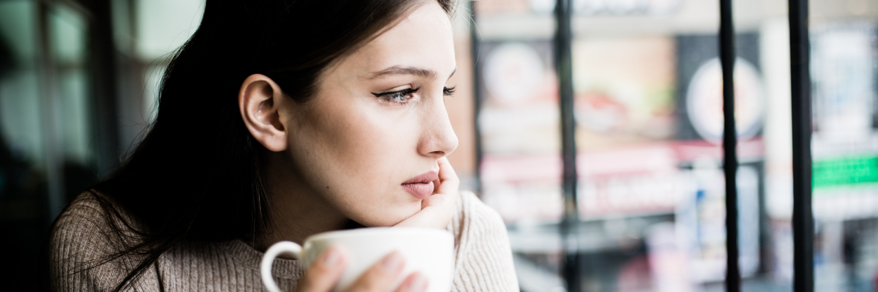 A picture of a woman sitting at a coffee shop, looking out the window with a sad expression.