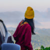 young woman in a yellow beanie and red jacket standing outside her car looking at the mountains