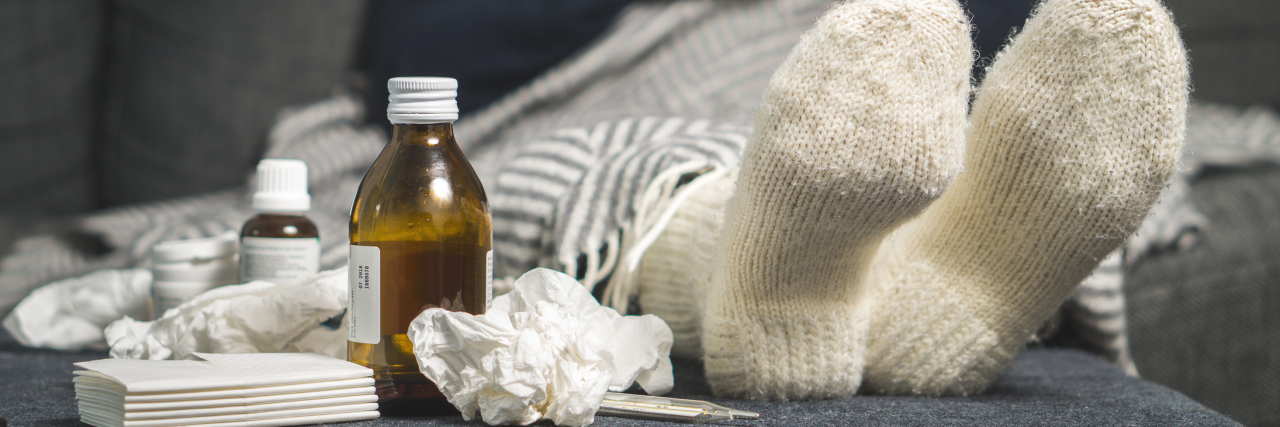 Why I Struggle More With Common Illnesses Due to My Chronic Illness woman sitting on the couch holding a mug of tea next to cough medicine and tissues