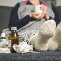 woman sitting on the couch holding a mug of tea next to cough medicine and tissues