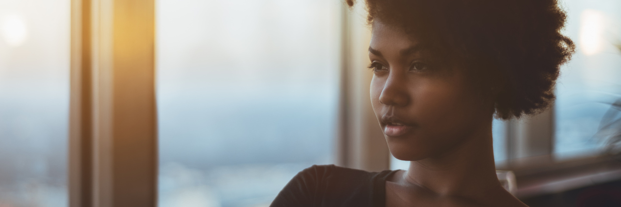 Why I'm Happy Even Though I Have Lupus, CRPS Portrait of pensive stately young female with curly hair sitting in dark settings next to big window, folds her arms and thinking about something; warm evening outside