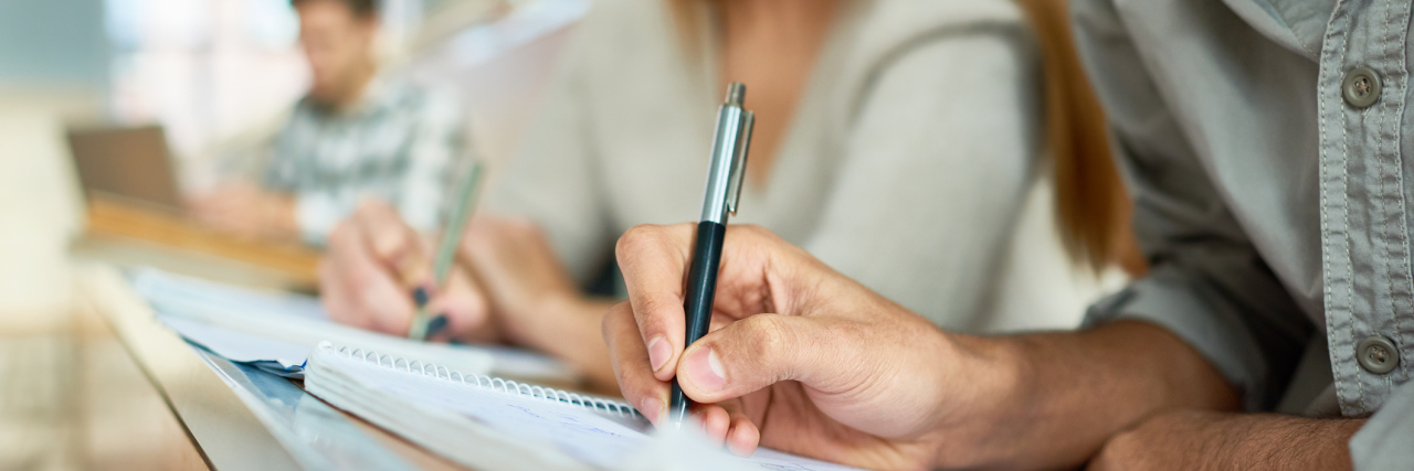 How Colleges Should Accommodate People with Chronic Illness Close up of male student taking notes sitting at desk during lecture in college, focus on hand writing in notebook, copy space
