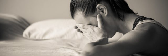 Stressed woman with hand over face leaning on bed.
