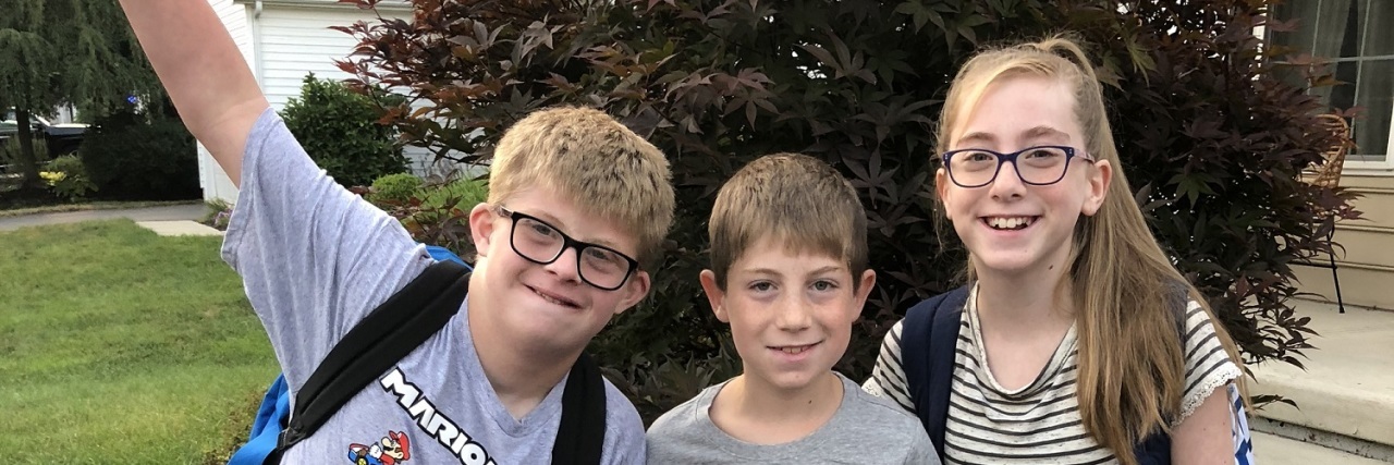It Takes an Inclusive Village to Raise Our Kids With Disabilities Three siblings posing forfirst day of school picture, one boy has Down syndrome