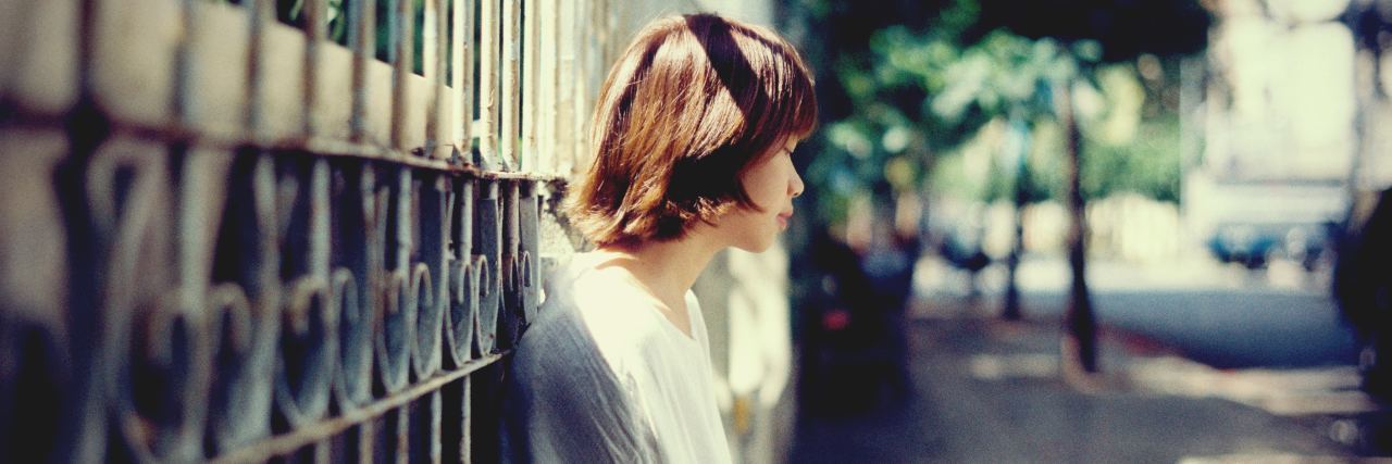 A photo of a woman outside, leaning on a gate, looking away from teh camera with a sad expression.