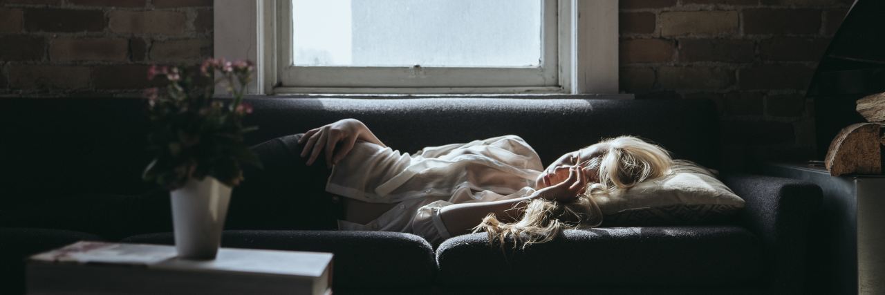 The Scary Way Fibromyalgia Pain Begins woman lying on her couch in a dark room during the day