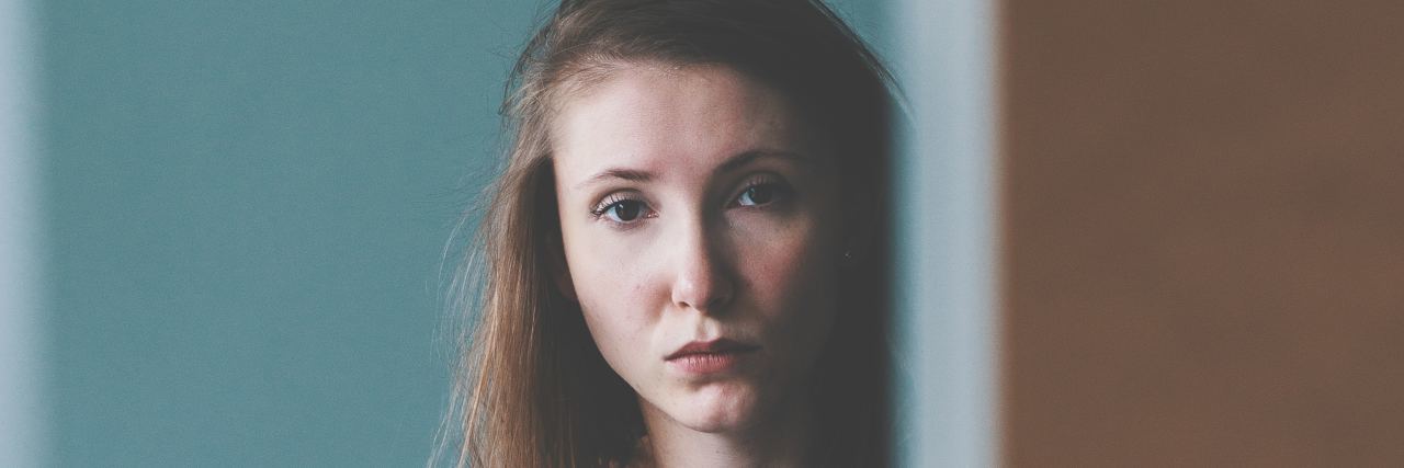 10 Feelings That Alternate in My Mind as I Live With ME/CFS A picture of a woman standing behind a wall, with a serious expression.