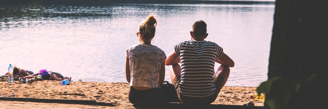 A picture of a young couple sitting on the ground outside, looking at a body of water.