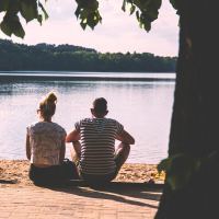 A picture of a young couple sitting on the ground outside, looking at a body of water.