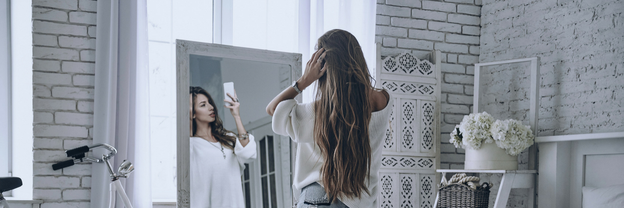 Plastic Surgeons Seeing More "Snapchat Dysmorphia," Body Dysmorphia Woman in a white shirt and shorts taking a selfie in a gray bedroom