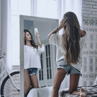 Woman in a white shirt and shorts taking a selfie in a gray bedroom