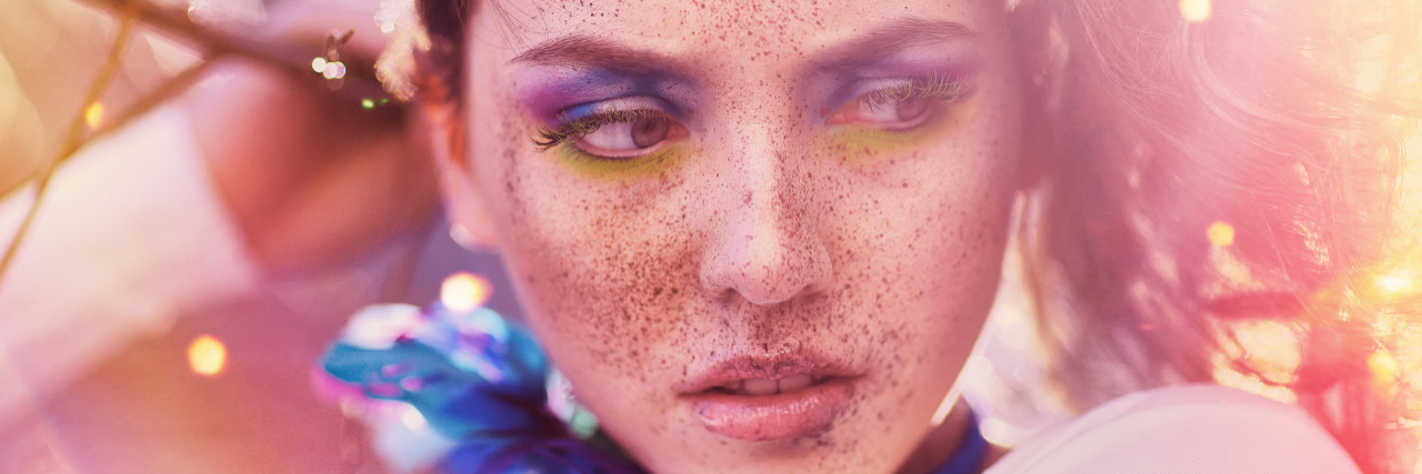 13 'Obsessive' Things People With Borderline Personality Disorder Do. A closeup of a young woman with freckles and bright blue and yellow eye makeup, the area around her blurry and bright with yellow sunlight. She's looking to the side with an ambiguous expression.