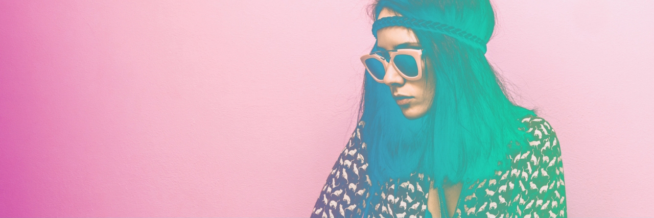 A woman with long hair, sunglasses, and a headband, in a style reminiscent of the 70s. The image is filtered to have vibrant colors, with the woman's hair appearing bright green, and she stands against a pink background. 19 Signs You Have POTS, Not 'Just' Anxiety