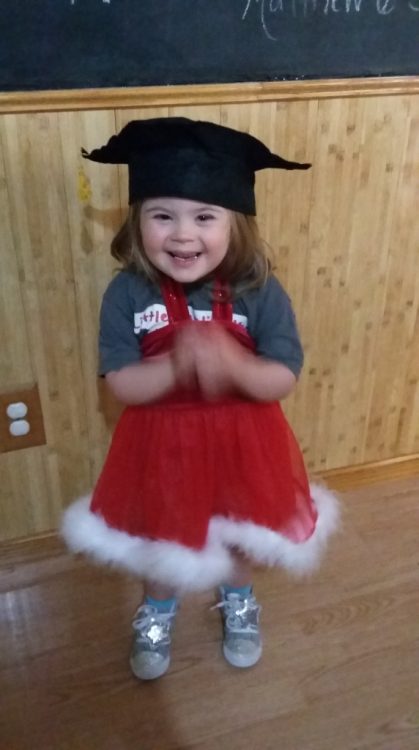 Little girl with Down syndrme wearing a graduation cap and a santa skirt while clapping her hands and smiling