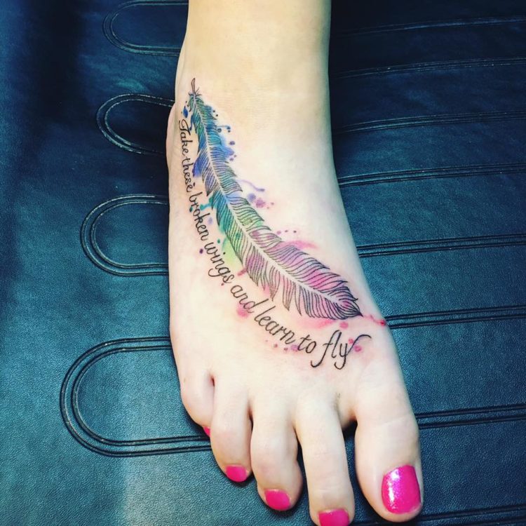 Tattoo of a multicolored feather on a foot. Beneath the feather reads, "Take these broken wings and learn to fly."