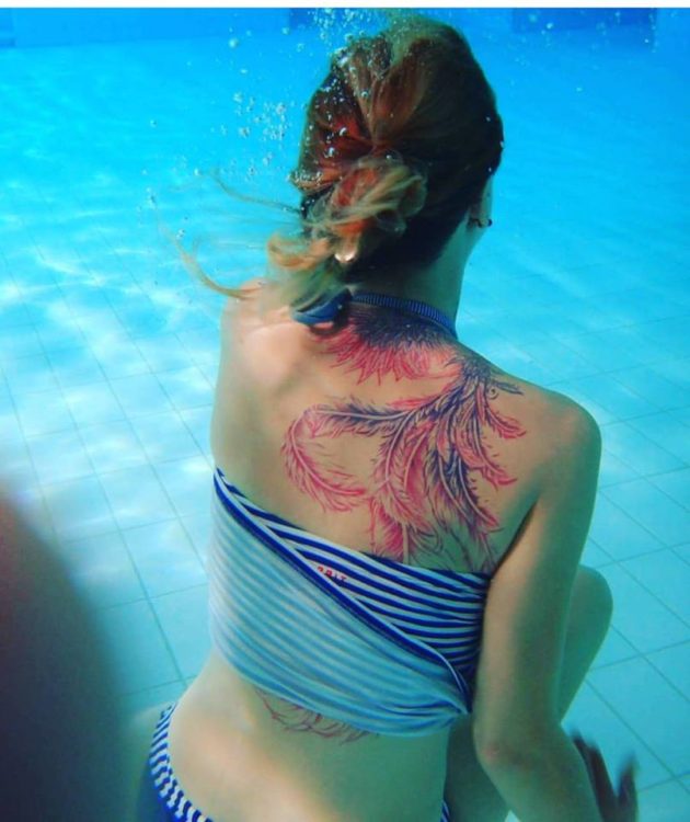 A woman in a swim suit sits in a pool facing away. A tattoo of blue and pink phoenix feathers wraps around the back of her neck and down her right shoulder blade.