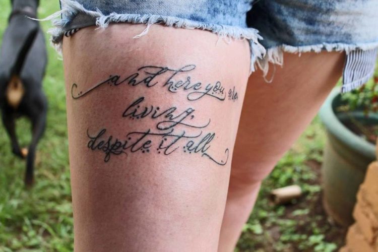 Tattoo on a thigh that reads: "and here you are living despite it all."