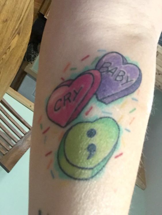 Tattoo on a forearm of three hearts. The pink heart reads "Cry", the purple heart reads "Baby", and the green heart has a semicolon.