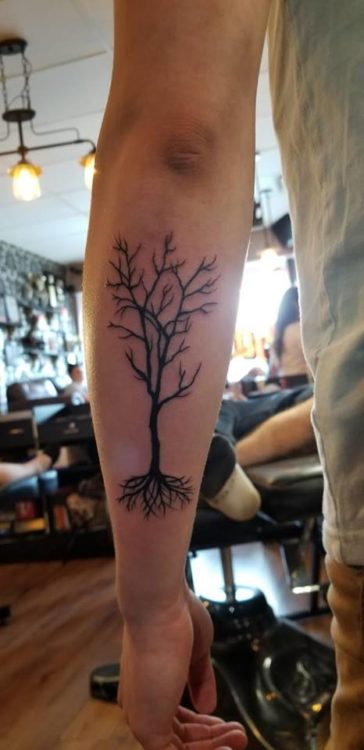 A man's forearm with a tattoo of a fire-blackened tree, with its branches bare and its roots exposed.