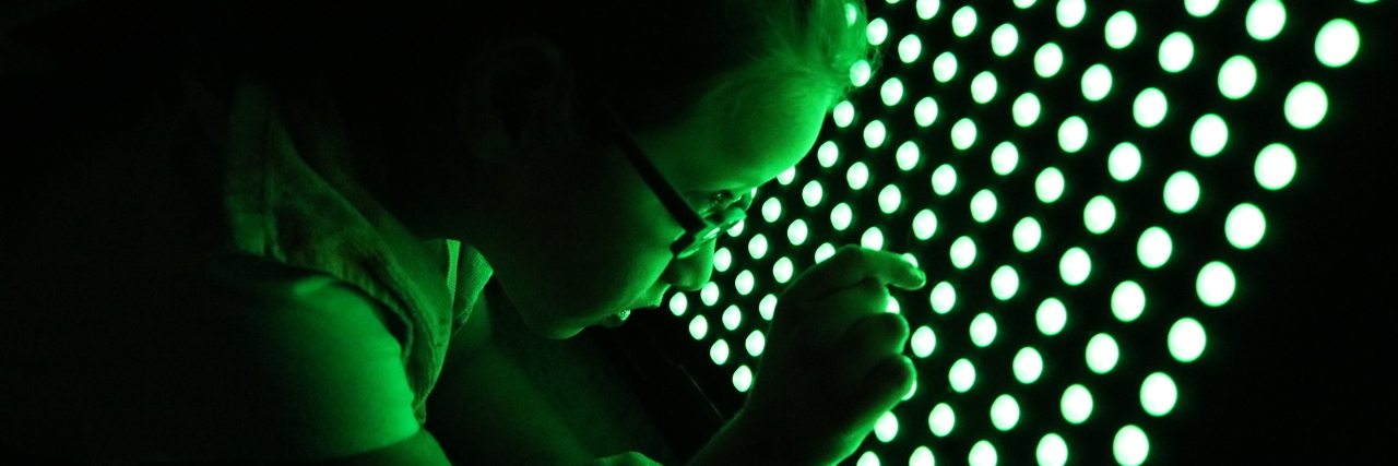 Little girl using a LightAide. It is a large board with green lights arranged in rows. She is putting her finger to one of the lights.