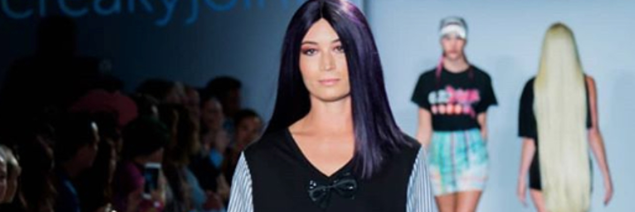 Charis Hill, a tall woman wearing a dark purple wig, walks down a runway.