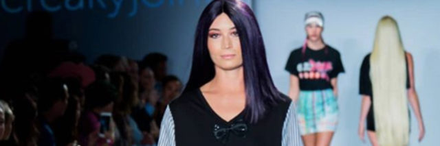 Charis Hill, a tall woman wearing a dark purple wig, walks down a runway.