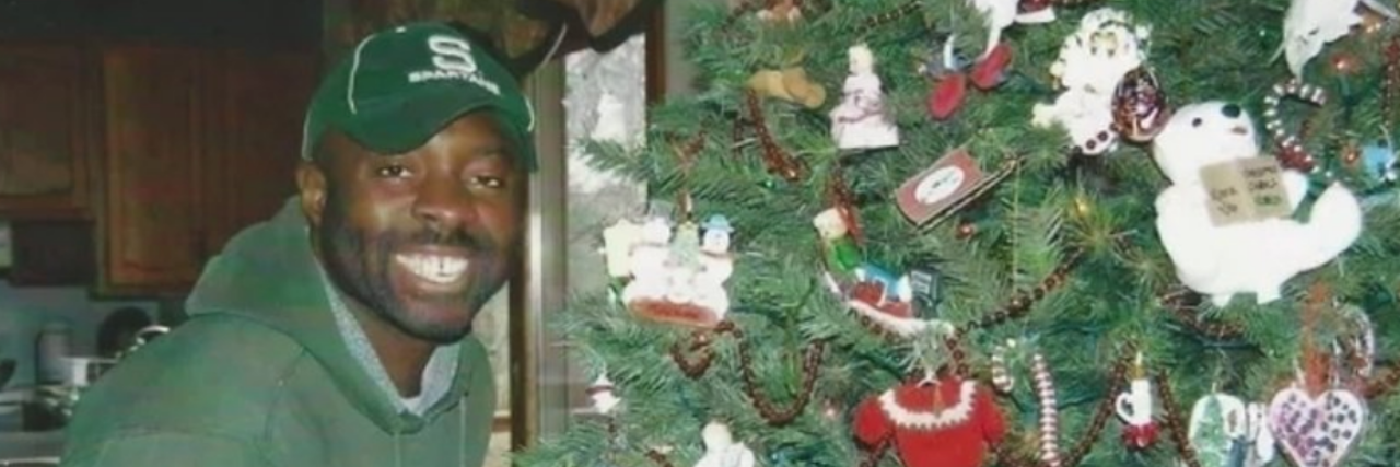 Francis Anwana standing next to a Christmas tree while wearing a green baseball cap and sweatshirt.