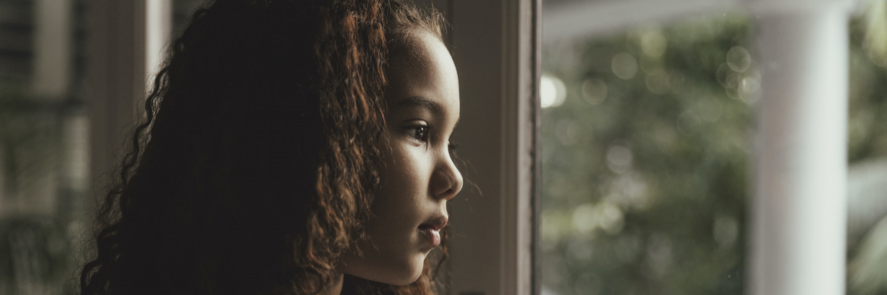 A young abused girl in a dim grey room looks out a window.