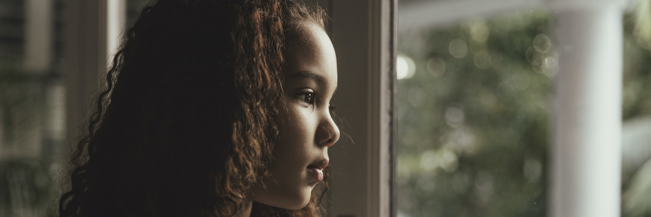 21 'Signs' of an Abusive Parent We Can't Keep Overlooking A young abused girl in a dim grey room looks out a window.
