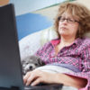 A tired looking woman sitting in bed in her pajamas, on her computer.