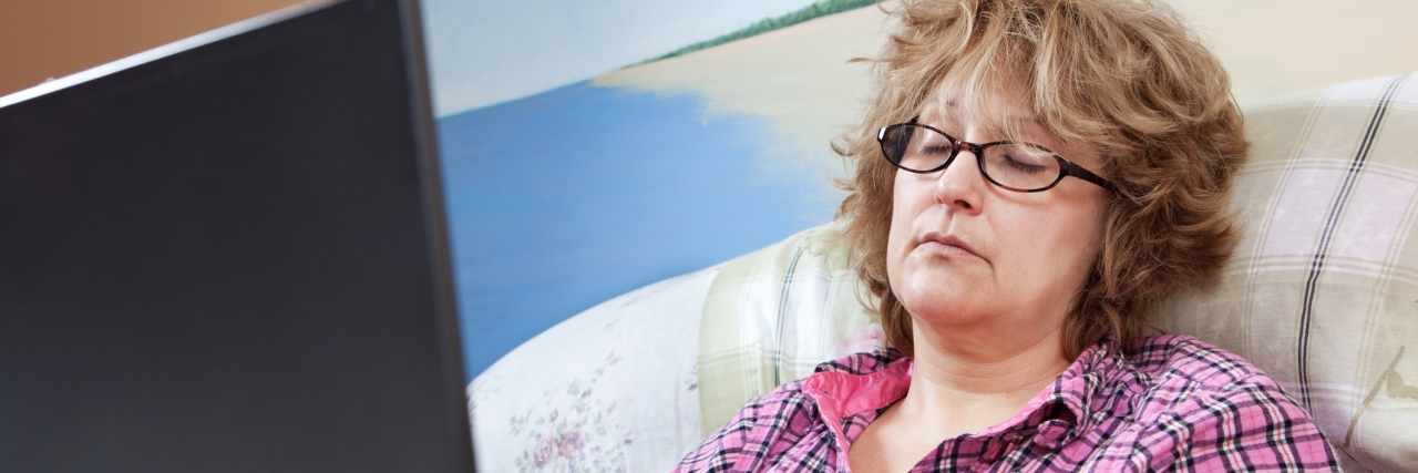 Pushing Too Hard With Fibromyalgia Means, 'If You Play, You Will Pay' A tired looking woman sitting in bed in her pajamas, on her computer.